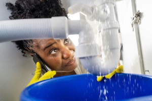 A woman talks on the phone while inspecting a leaking sink pipe with water issues. The concept is to avoid water leaks.